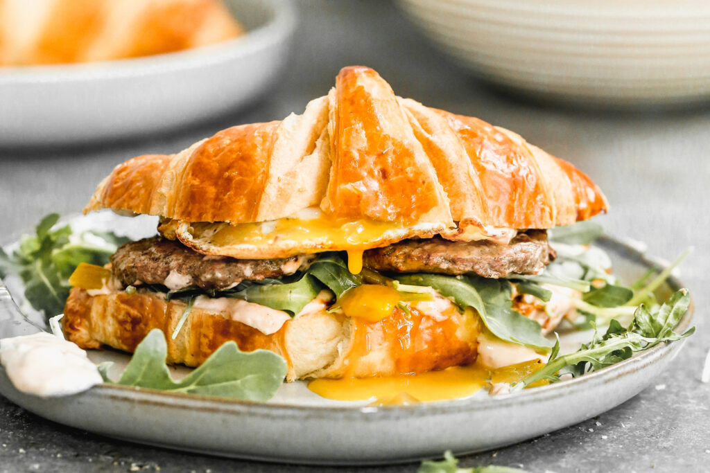 A breakfast sandwich with egg, sausage, and arugula on a croissant.