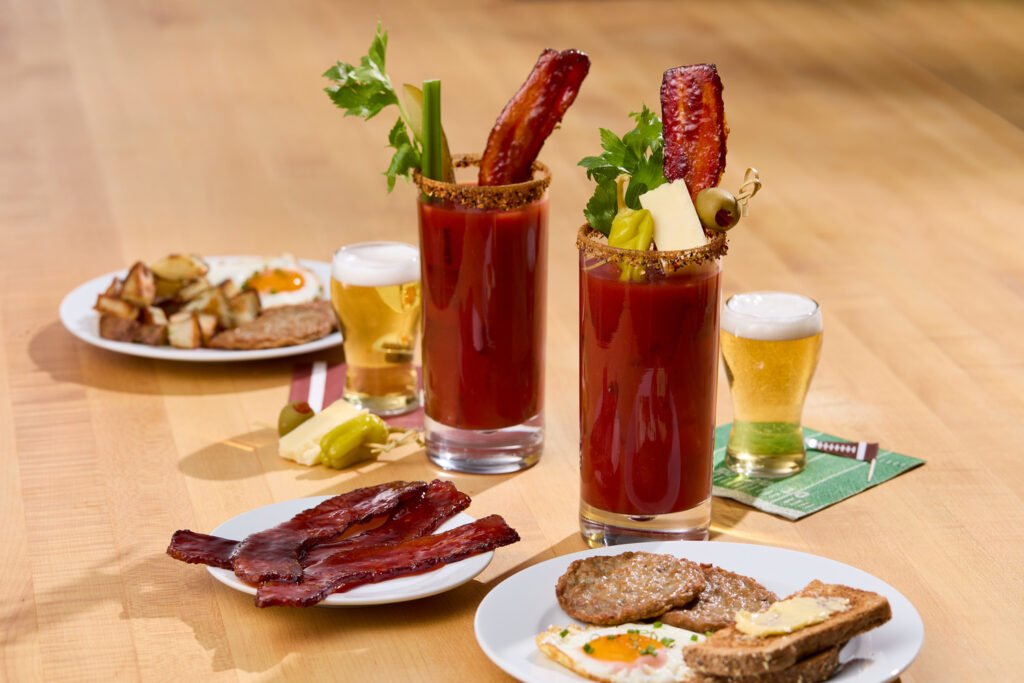 A plate of candied bacon in front of two Bloody Marys with Millionaire’s Bacon