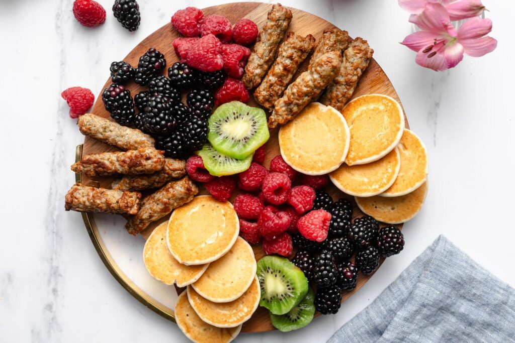 A breakfast board with Jones Dairy Farm chicken sausage, mini pancakes, raspberries, kiwi, and blackberries.